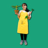 mood shot of  girl wearing Daily Dump Compost Apron in outdoor garden and holding rake from gardening toolkit and potted plant in the other