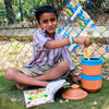 Young smiling boy seated outdoors reading and disposing banana peel into Daily Dump Nano khamba by his side.